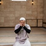 Muslim man in a white prayer cap kneels on a beige patterned carpet hands covering his face in prayer inside a spacious hall