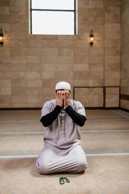 Muslim man in a white prayer cap kneels on a beige patterned carpet hands covering his face in prayer inside a spacious hall