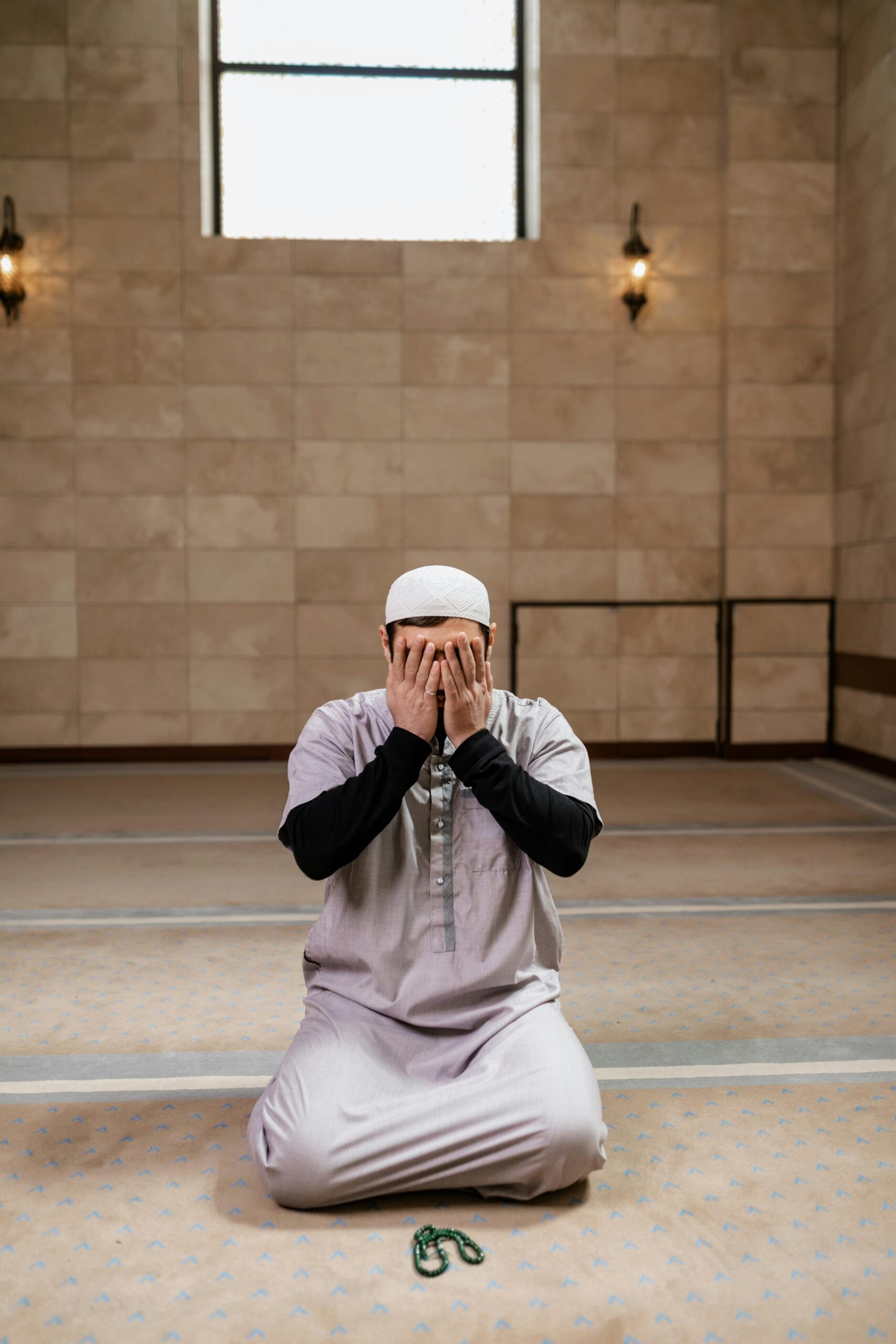 Muslim man in a white prayer cap kneels on a beige patterned carpet hands covering his face in prayer inside a spacious hall