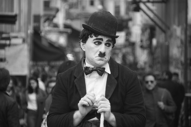 Mime street performer with white face makeup, bowler hat, dark suit, and cane in a crowded city street.