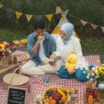 Two people sit on a red and white checked picnic blanket with snacks flowers and bunting in a garden setting