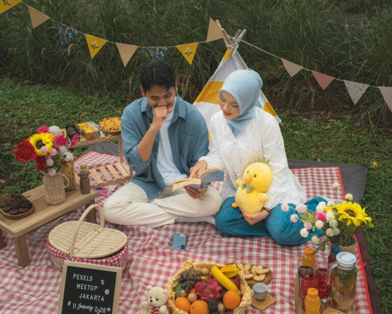 Two people sit on a red-and-white checked picnic blanket with snacks, flowers and bunting in a garden setting.