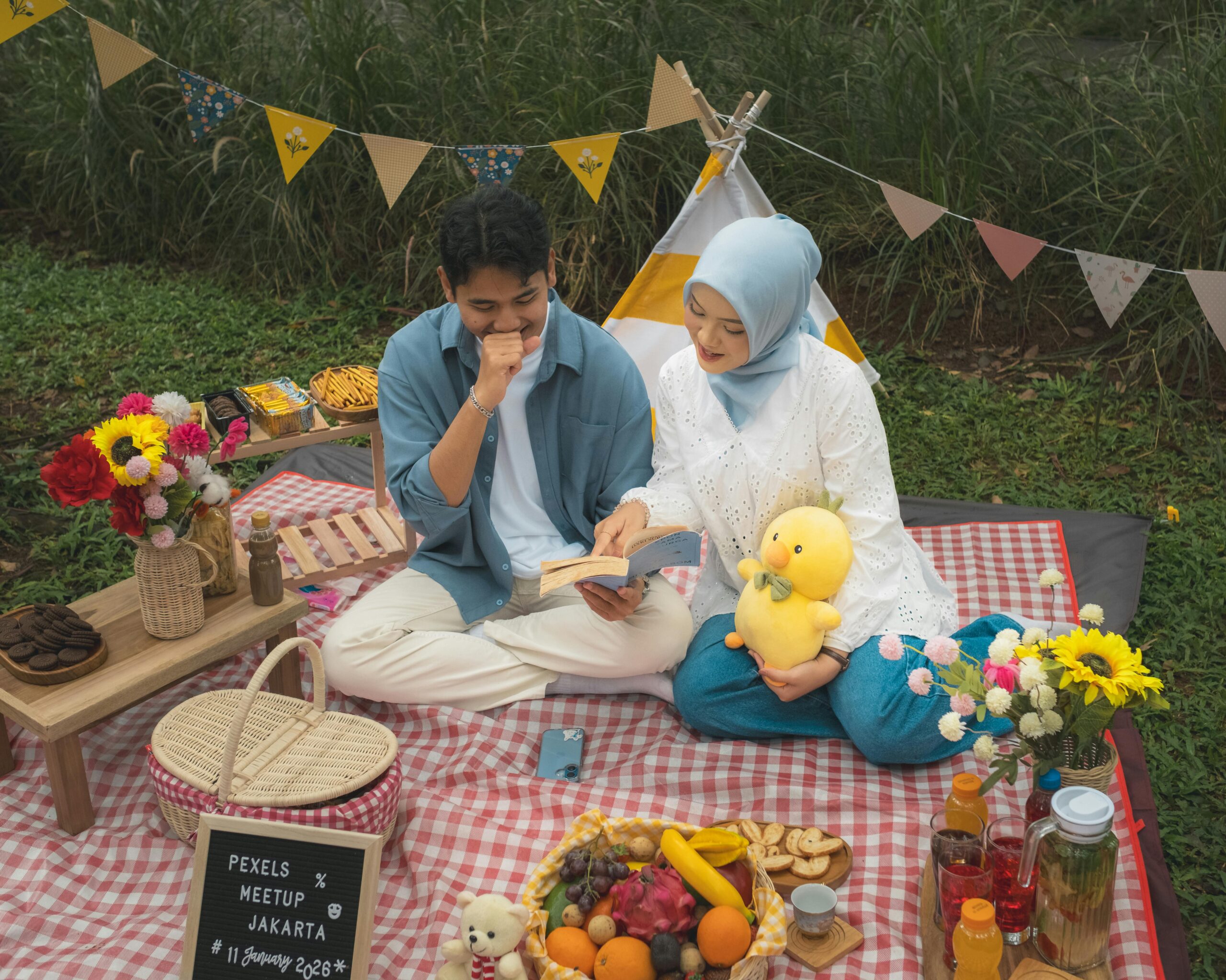 Two people sit on a red-and-white checked picnic blanket with snacks, flowers and bunting in a garden setting.