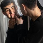 Young man in a black sweatshirt presses his lips with his fingers while looking at his reflection in a mirror