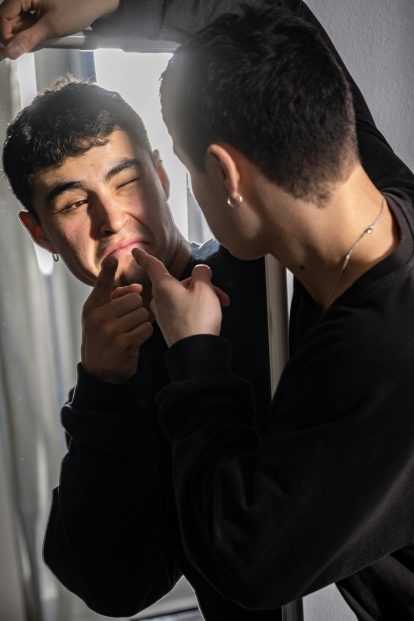 Young man in a black sweatshirt presses his lips with his fingers while looking at his reflection in a mirror.