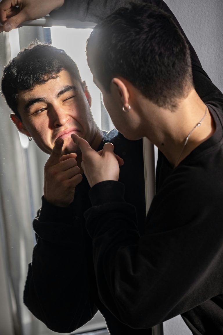 Young man in a black sweatshirt presses his lips with his fingers while looking at his reflection in a mirror.