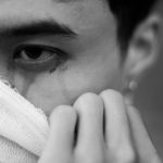 Close up of a person wearing a torn bandage over the mouth glancing sideways with a tear on the cheek black and white image conveying injury or distress