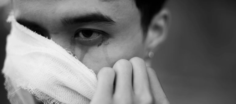 Close-up of a person wearing a torn bandage over the mouth, glancing sideways with a tear on the cheek; black-and-white image conveying injury or distress.