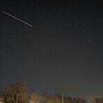 Night sky filled with stars over a lit parking lot and leafless trees a diagonal light streak crosses the left side of the sky