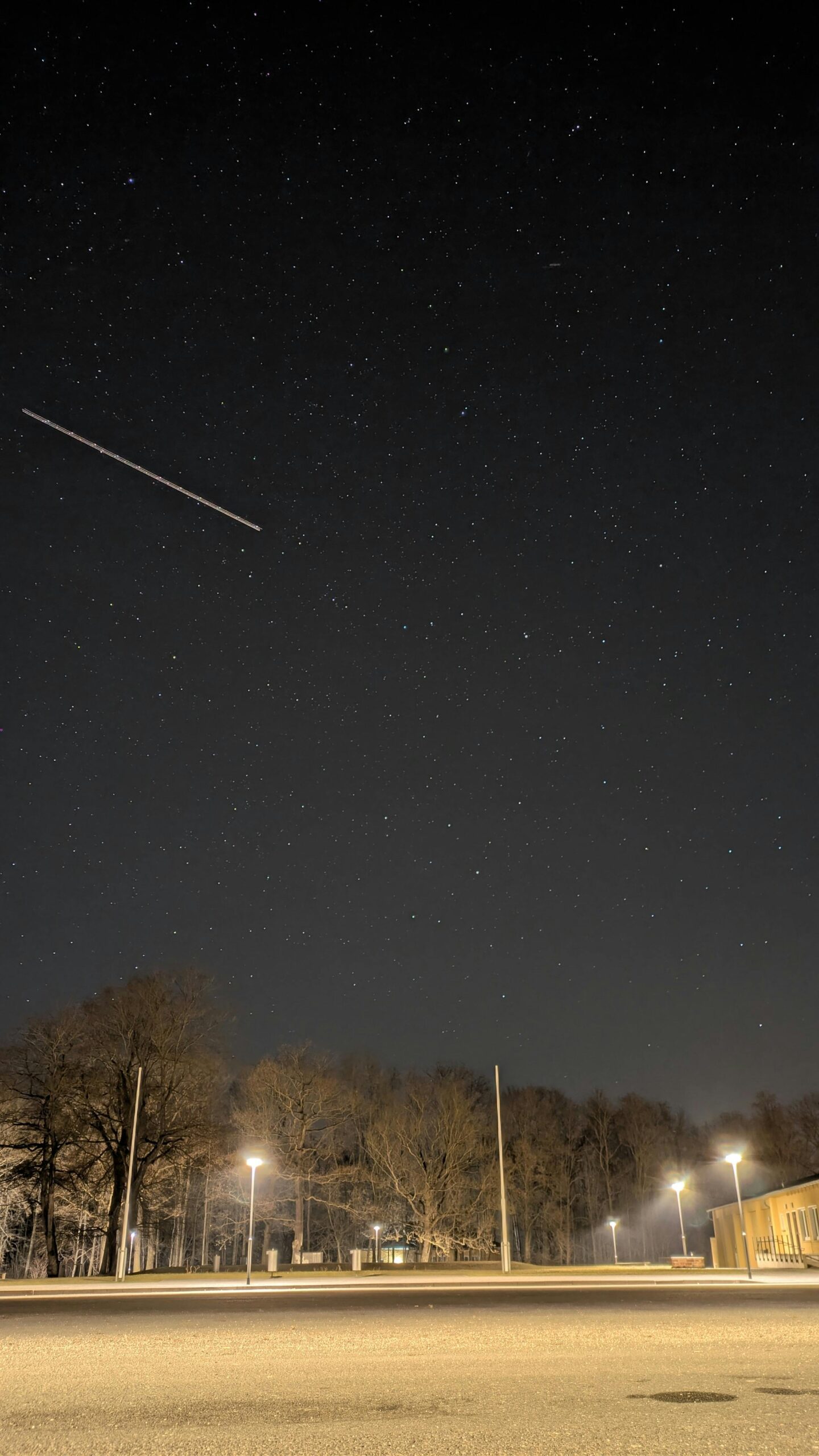 Pexelsmanuelbluemel215912686337014202 catrawarta Night sky filled with stars over a lit parking lot and leafless trees a diagonal light streak crosses the left side of the sky