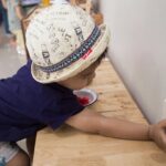 Child in a printed hat reaches to insert or touch a wall outlet mounted on a wooden counter