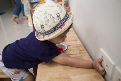 Child in a printed hat reaches to insert or touch a wall outlet mounted on a wooden counter.