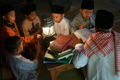 Children gathered around a lit lantern studying from stacked religious books on a wooden table in a dim room.