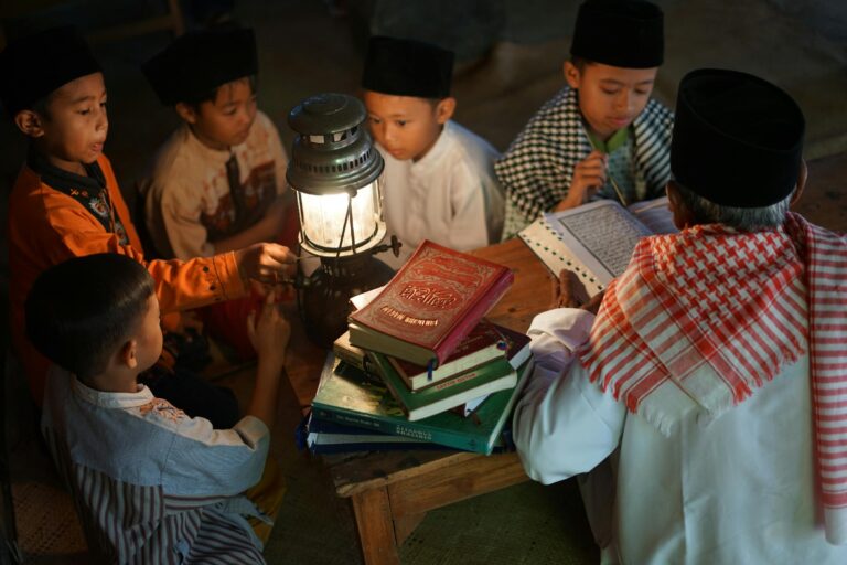 Children gathered around a lit lantern studying from stacked religious books on a wooden table in a dim room.