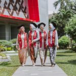 Four women in hijabs and graduation gowns walk together outside a modern campus building wearing red stoles with 2020