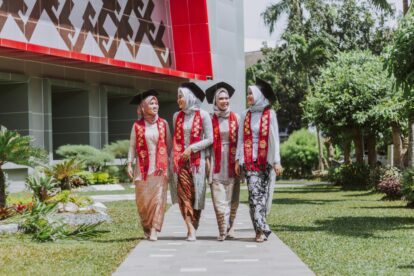 Four women in hijabs and graduation gowns walk together outside a modern campus building, wearing red stoles with 2020.