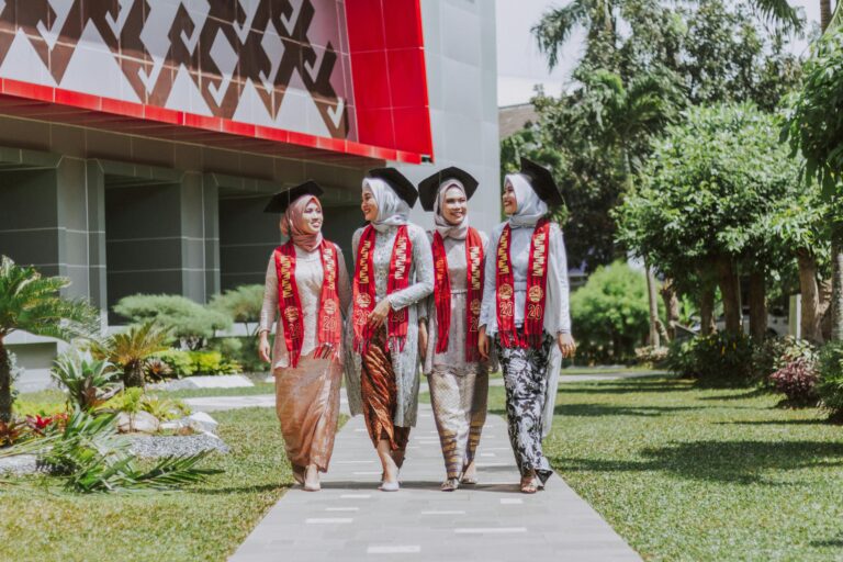 Four women in hijabs and graduation gowns walk together outside a modern campus building, wearing red stoles with 2020.