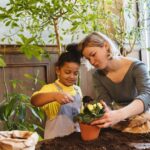 A woman helps a young girl plant flowers in a small terracotta pot indoors with soil on the table and greenery around them