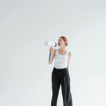 Woman in a white tank top and black pants shouting into a megaphone in a bright empty space