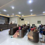 Waiting area in a government service office with brown and tan couches and people seated desks and staff at the far counter sign reading samsat pembantu sewon bantul on the wall in the background