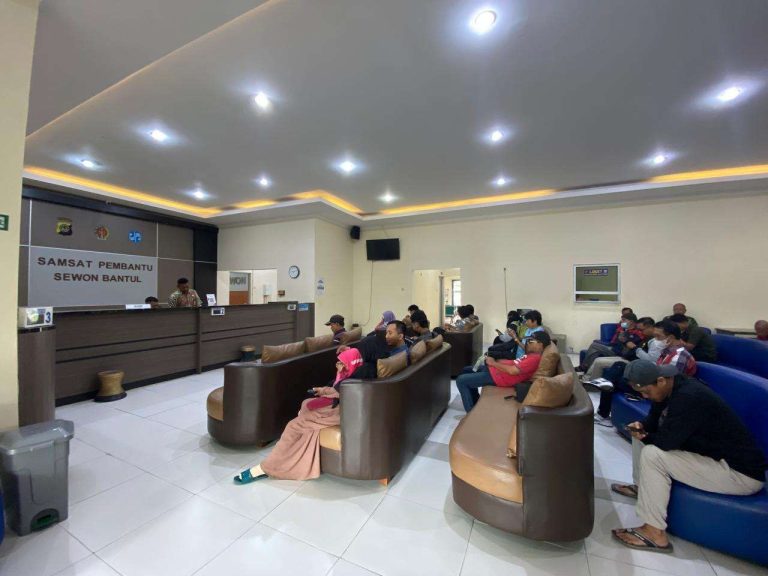Waiting area in a government service office with brown and tan couches and people seated, desks and staff at the far counter, sign reading 'SAMSAT Pembantu Sewon Bantul' on the wall in the background.