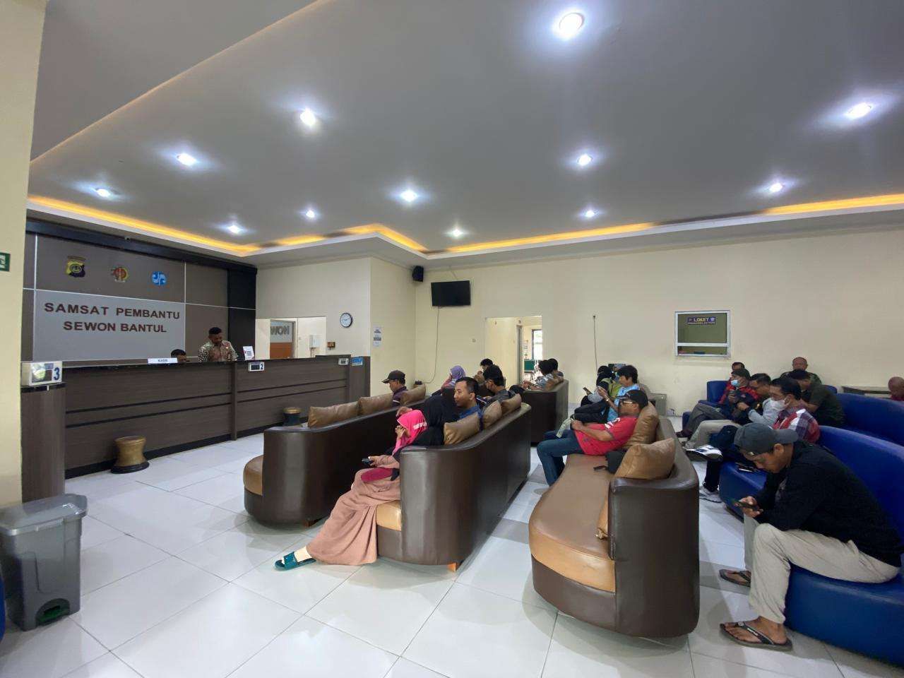 Waiting area in a government service office with brown and tan couches and people seated desks and staff at the far counter sign reading samsat pembantu sewon bantul on the wall in the background
