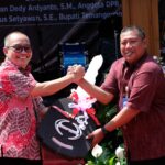 Two men on a stage shake hands and smile while presenting a black plaque with pink ribbons in the foreground