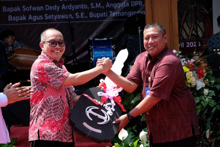 Two men on a stage shake hands and smile while presenting a black plaque with pink ribbons in the foreground.