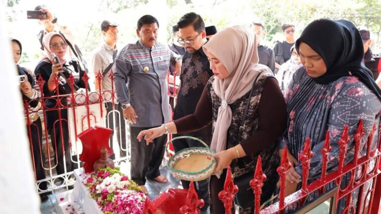 Group of people, mainly women in hijabs, placing flowers and petals at a shrine behind a red fence while others observe.