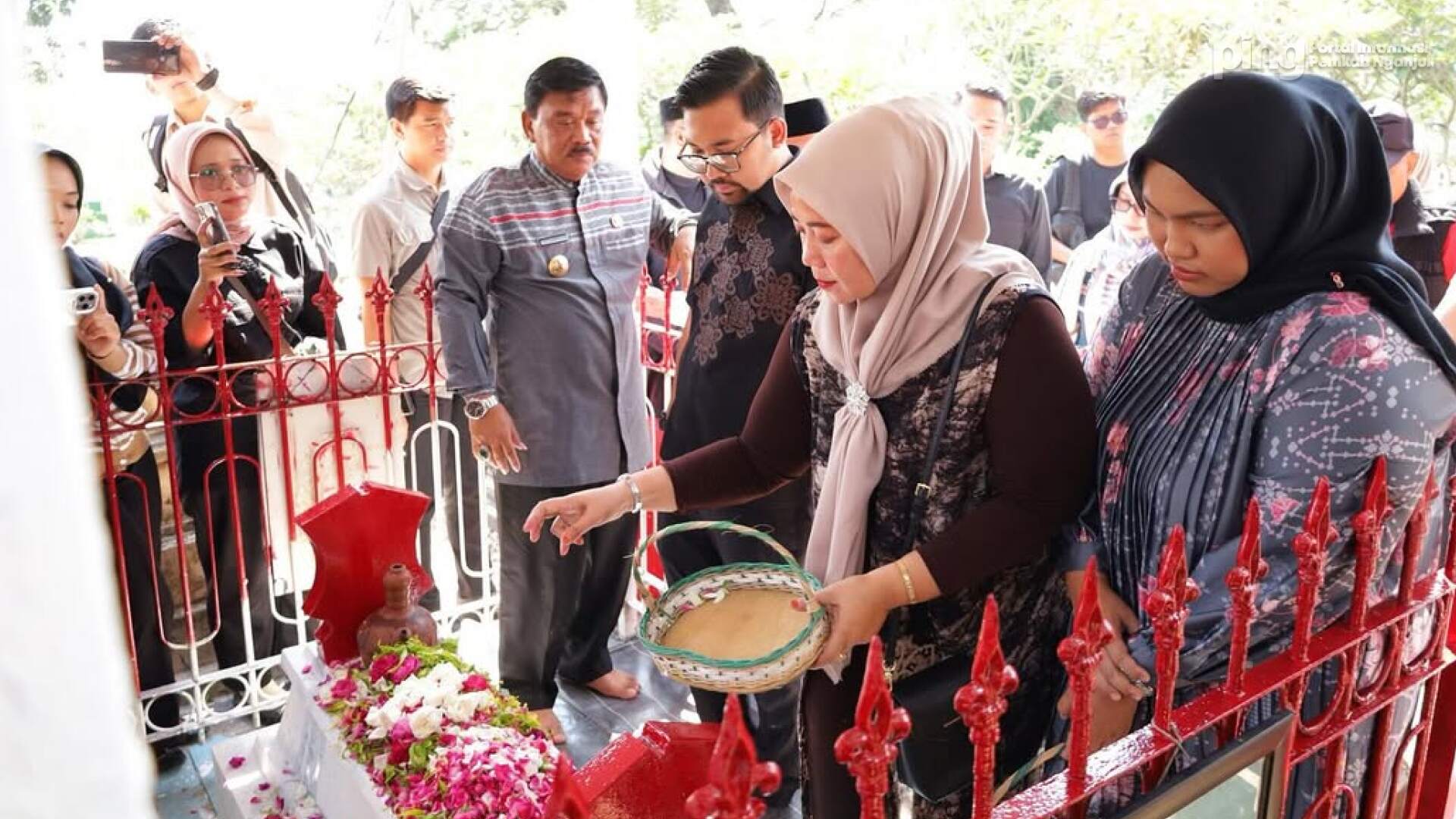 Group of people mainly women in hijabs placing flowers and petals at a shrine behind a red fence while others observe