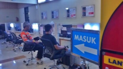 Waiting area at a service counter in a public office two people sit facing desks with a blue masuk sign and a left arrow nearby