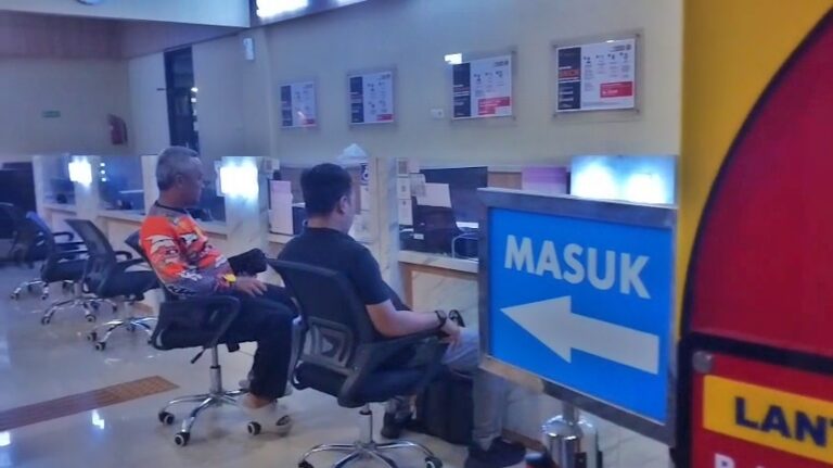 Waiting area at a service counter in a public office; two people sit facing desks, with a blue MASUK sign and a left arrow nearby.