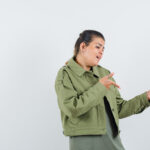 Woman in an olive green jacket gesturing with her hands as if explaining something against a plain light background