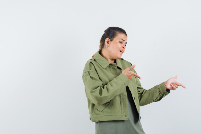 Woman in an olive-green jacket gesturing with her hands as if explaining something against a plain light background