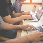 Multiple people typing on laptops at a shared desk in a bright office sunlight from a window in the background