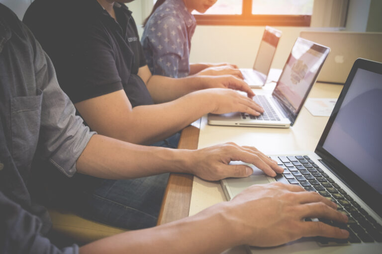 Multiple people typing on laptops at a shared desk in a bright office, sunlight from a window in the background.