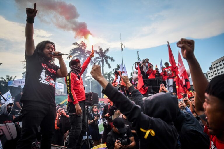 Male singer with microphone on stage at a protest, crowd raising fists and red flags; pink smoke in the sky behind him.