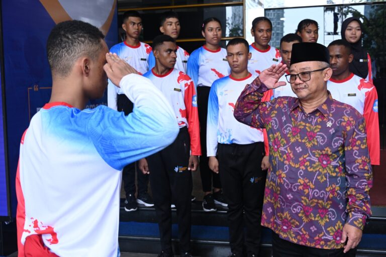 Group of students in matching white-and-blue jerseys salute a man in a patterned batik shirt and black cap during a ceremony, outdoors.