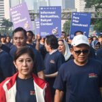 Group of people at a rally wearing navy shirts holding large purple and blue signs with indonesian text in an urban setting