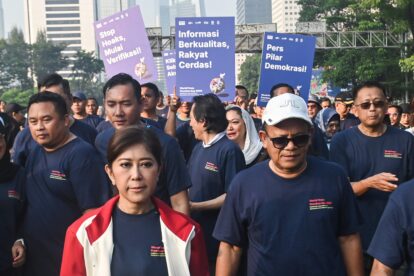 Group of people at a rally wearing navy shirts, holding large purple and blue signs with Indonesian text in an urban setting.
