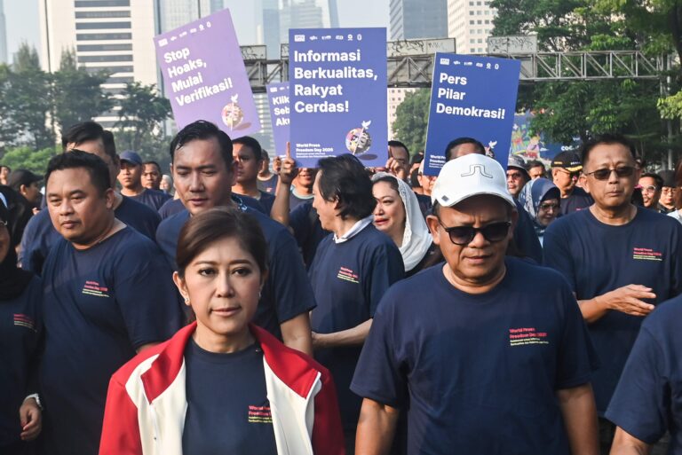 Group of people at a rally wearing navy shirts, holding large purple and blue signs with Indonesian text in an urban setting.