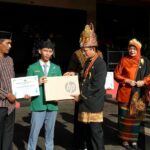 Student in a green blazer receives a prize from a man in traditional attire on a stage holding an hp box and a certificate nearby