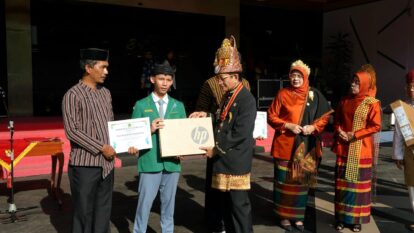 Student in a green blazer receives a prize from a man in traditional attire on a stage, holding an HP box and a certificate nearby.