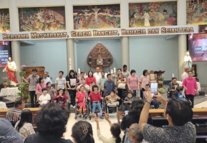 Group of people on a church stage including several children in wheelchairs posing for a photo under a banner audience takes pictures from the pews