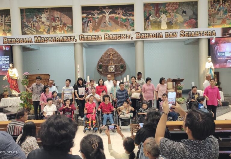 Group of people on a church stage, including several children in wheelchairs, posing for a photo under a banner; audience takes pictures from the pews.