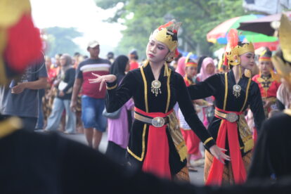 Group of performers in ornate black and red traditional costumes with gold headdresses dancing in a street parade, with a crowd watching in the background.