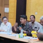 Panel of five people seated at a conference table one man in batik and headscarf speaks into a microphone while others listen water bottles and yellow snack boxes on the table in a meeting room