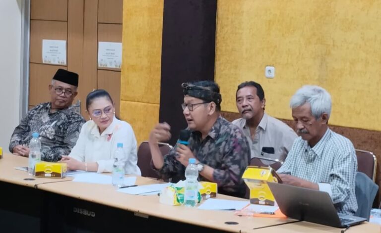 Panel of five people seated at a conference table, one man in batik and headscarf speaks into a microphone while others listen, water bottles and yellow snack boxes on the table in a meeting room.