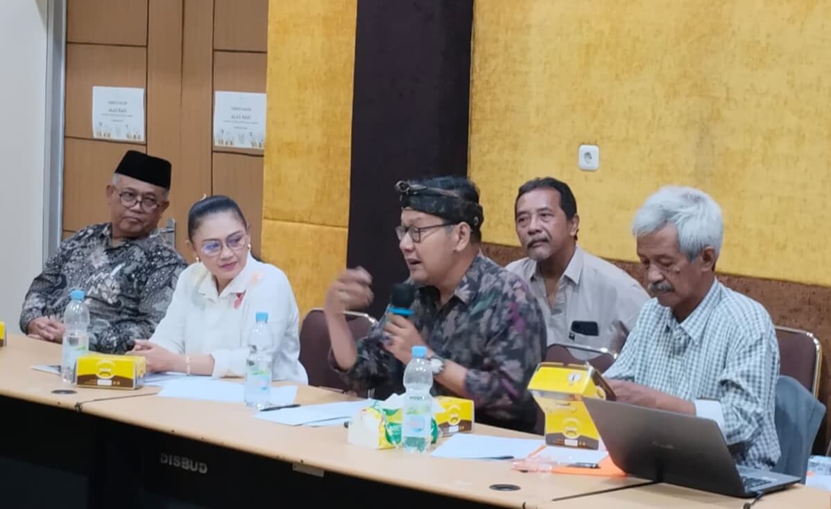 05dahlan 1 catrawarta Panel of five people seated at a conference table one man in batik and headscarf speaks into a microphone while others listen water bottles and yellow snack boxes on the table in a meeting room
