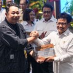 Two men shake hands while presenting a glass award plaque the recipient in a white uniform smiles as onlookers watch in the background